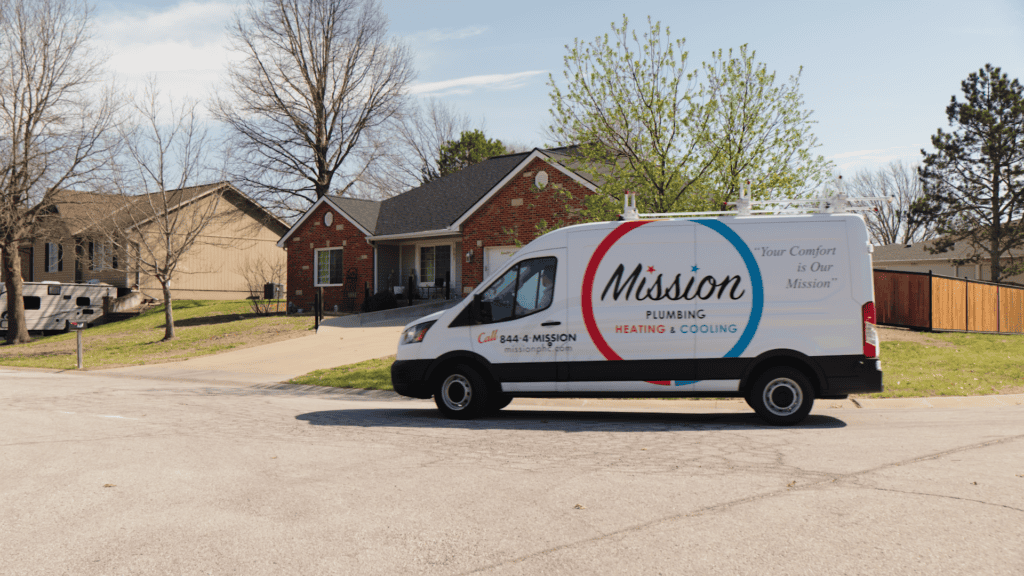 A Mission Plumbing Heating & Cooling van is parked on a residential street in front of a brick house with trees and a driveway visible.