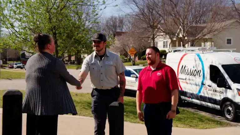 Two men, one in a uniform and another in a red shirt, stand outside as the man in uniform makes contact with a woman by shaking her hand. A Mission company van is parked in the background.