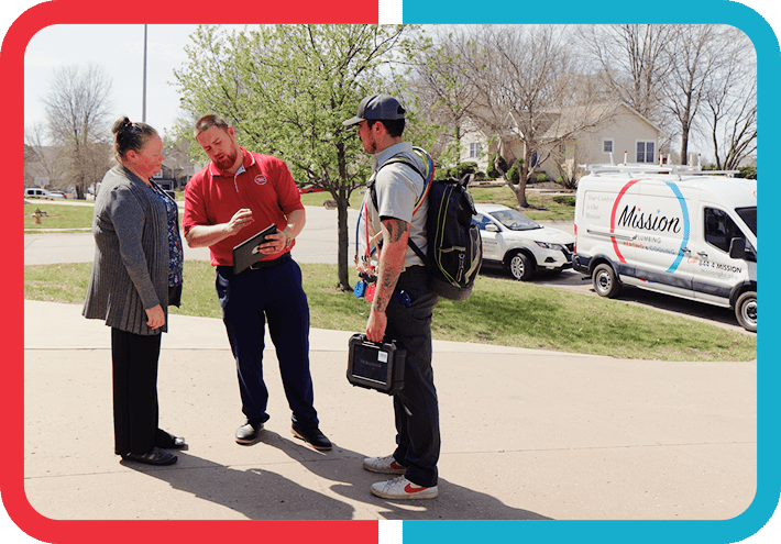 Three people stand talking outside near a Mission-branded van; one person holds a clipboard, another holds a case and wears a backpack.